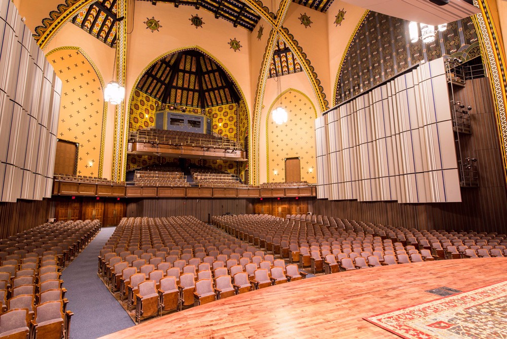 A shot of the irvine Main Hall seating area taken from its stage proscenium. The shot shows the entirety of the 1260 seat theartre's orchestra and balonies under beautiful stone arches painted in a yellow mosaic.