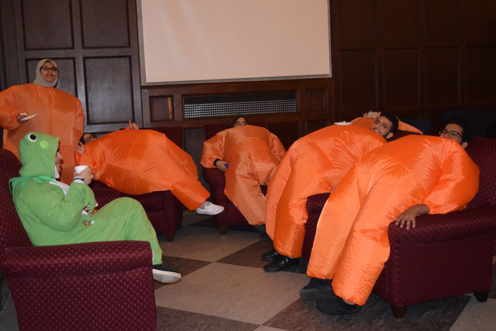 5 students in blow up orange haunt costumes comically posing in a lounge chair around one student in a frog onesie pajama costume