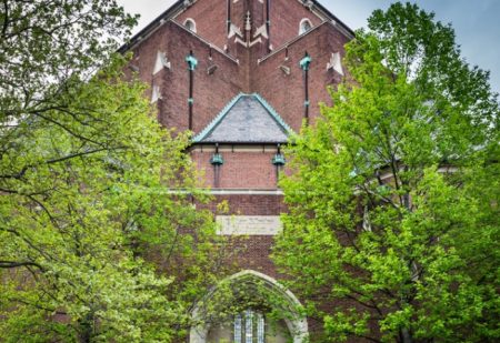 Exterior shot of Irvine Auditorium showing its red brick facade and church-like steeple. The building is framed by two lush green trees.