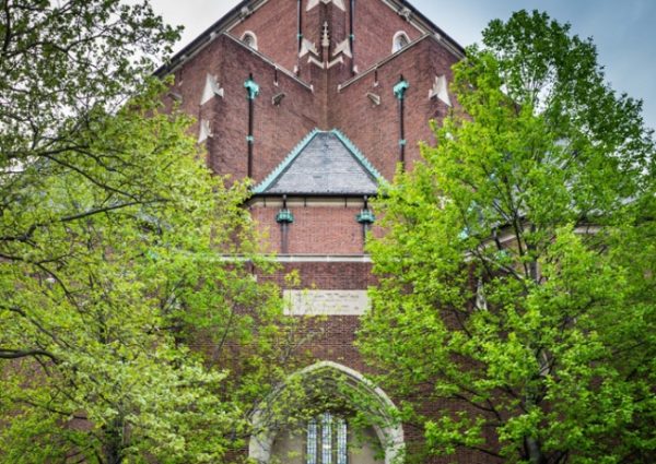Exterior shot of Irvine Auditorium showing its red brick facade and church-like steeple. The building is framed by two lush green trees.